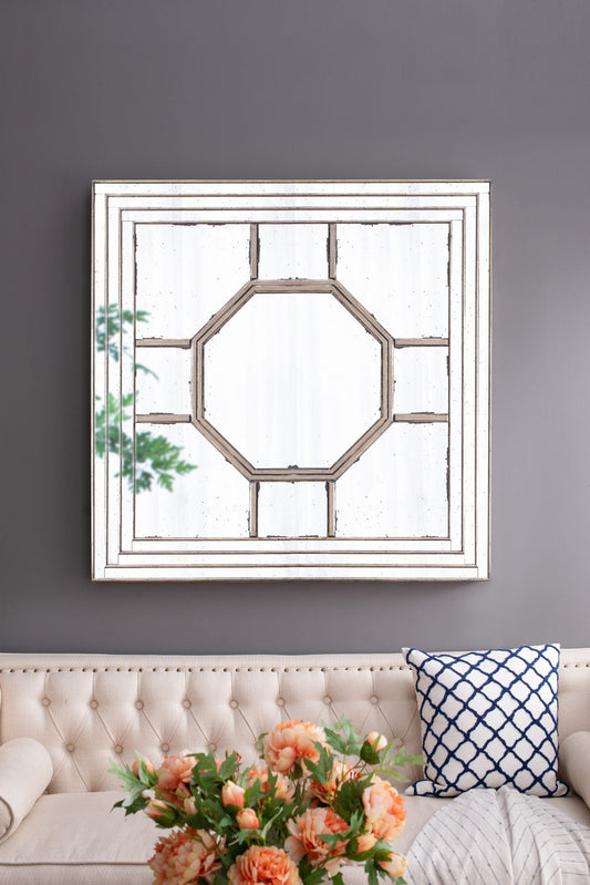 An Antique Style Decorative Square Wall Mirror with a mirrored frame hangs above a beige tufted sofa topped with peach flowers in a vase and a blue lattice cushion, reflecting a leafy branch in the living room.