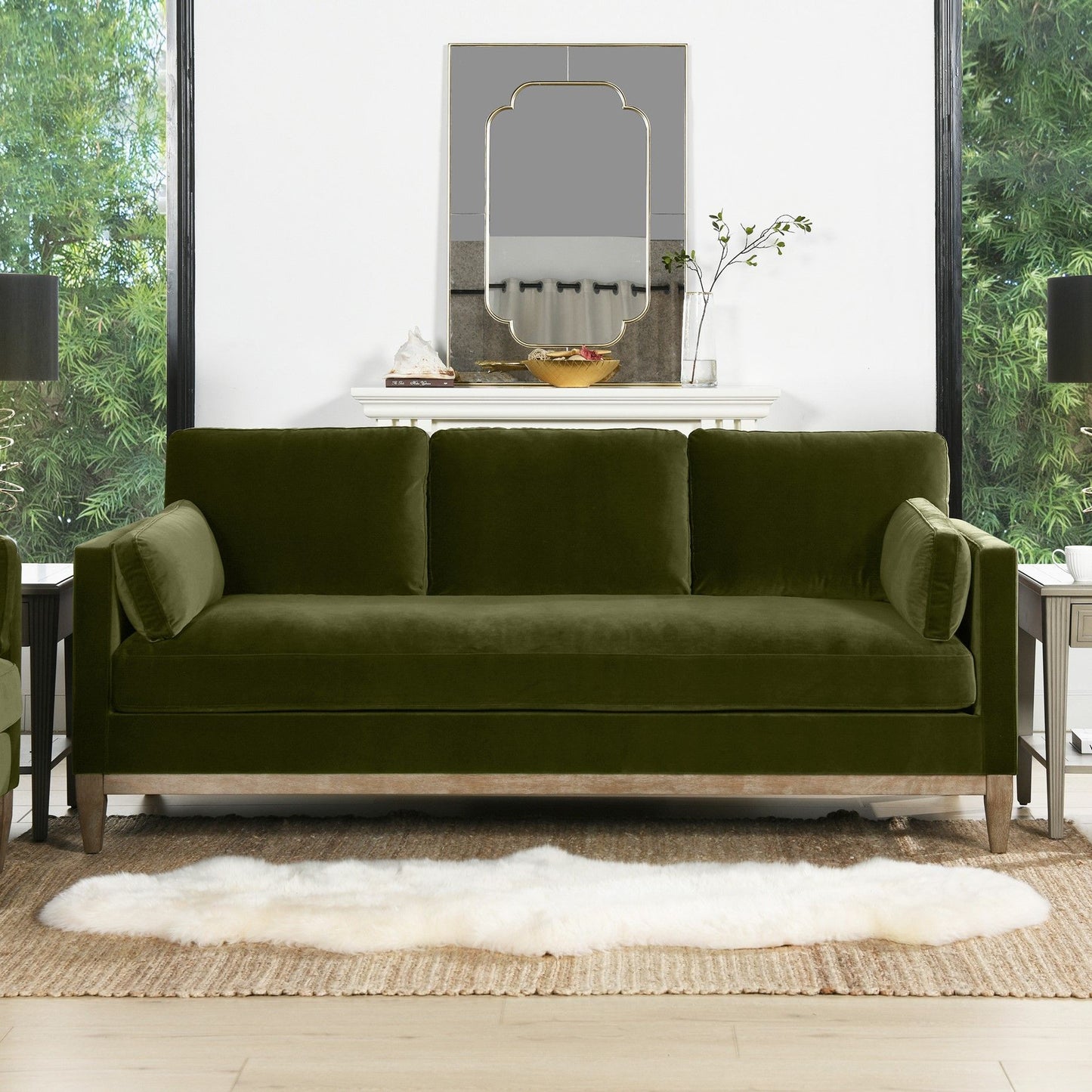 The Knox - Modern Farmhouse Sofa in green velvet, featuring three back cushions and two bolster pillows, sits on a white rug in a modern living room with decor, potted plant, and mirror on the mantle. Large windows reveal greenery outside.