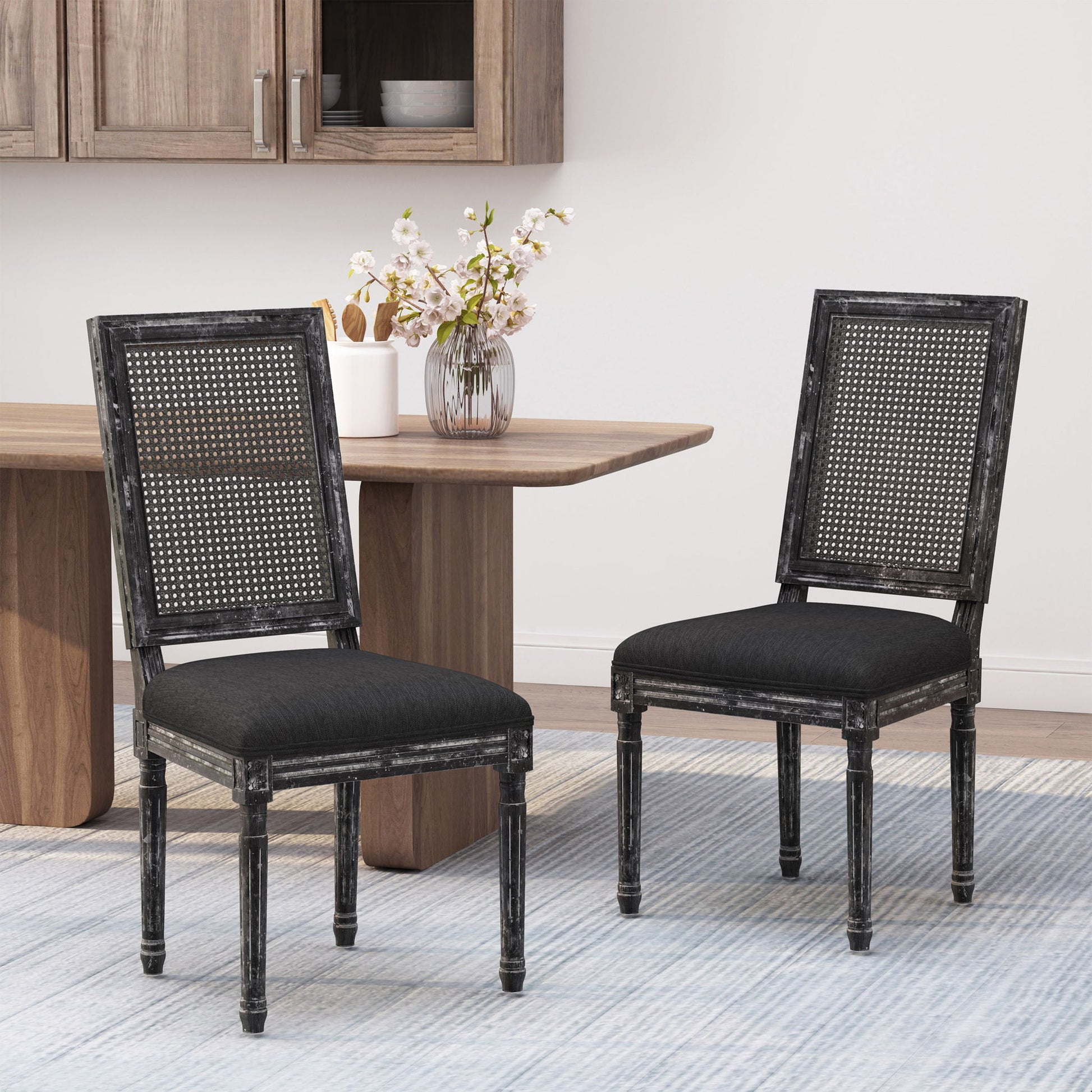 Two French Country Dining Chairs with rectangular cane backrests and smooth upholstered seats in black wood sit on a light blue striped rug in a modern dining room, beside a wooden table topped with a vase of flowers.