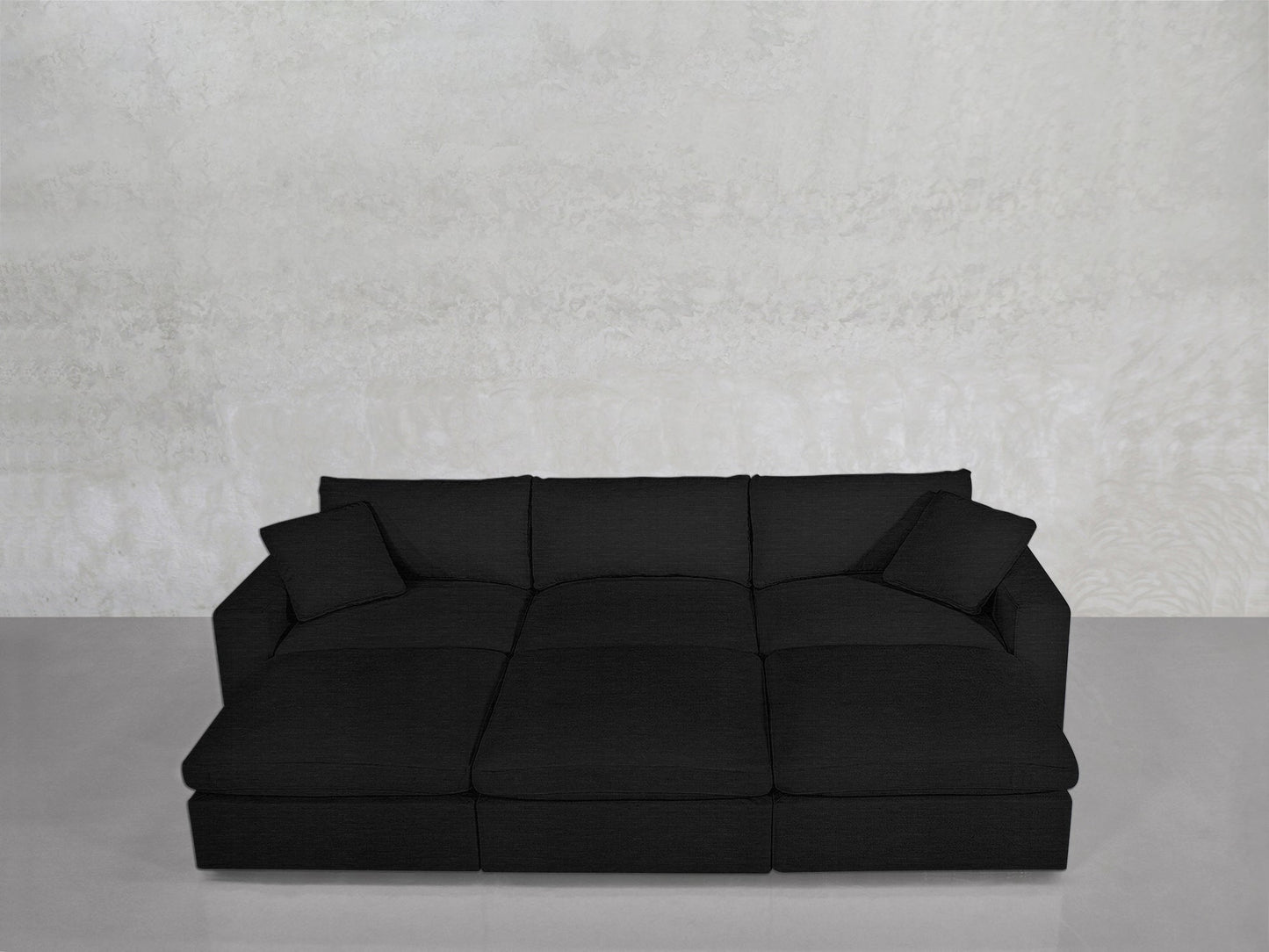 A sleek, black 6-Seat Modular Daybed with square armrests and two matching cushions sits against a light gray textured wall on a gray floor.