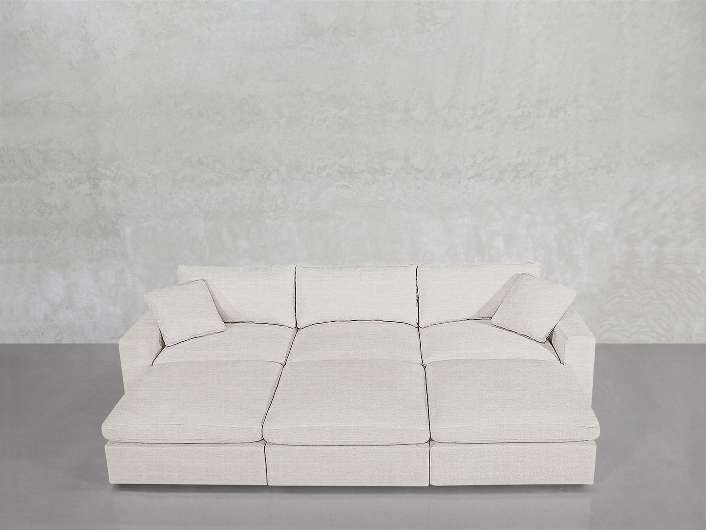 A contemporary 6-Seat Modular Daybed in light beige with three seat cushions and two matching throw pillows sits against a textured light gray wall on a polished gray floor.