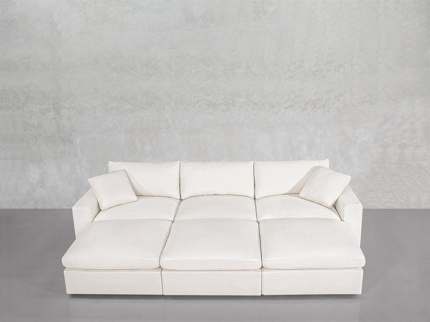 A modern, minimalist 6-Seat Modular Daybed in white, featuring three seat cushions, three back cushions, and two matching throw pillows, sits against a light gray textured wall on a smooth gray floor.