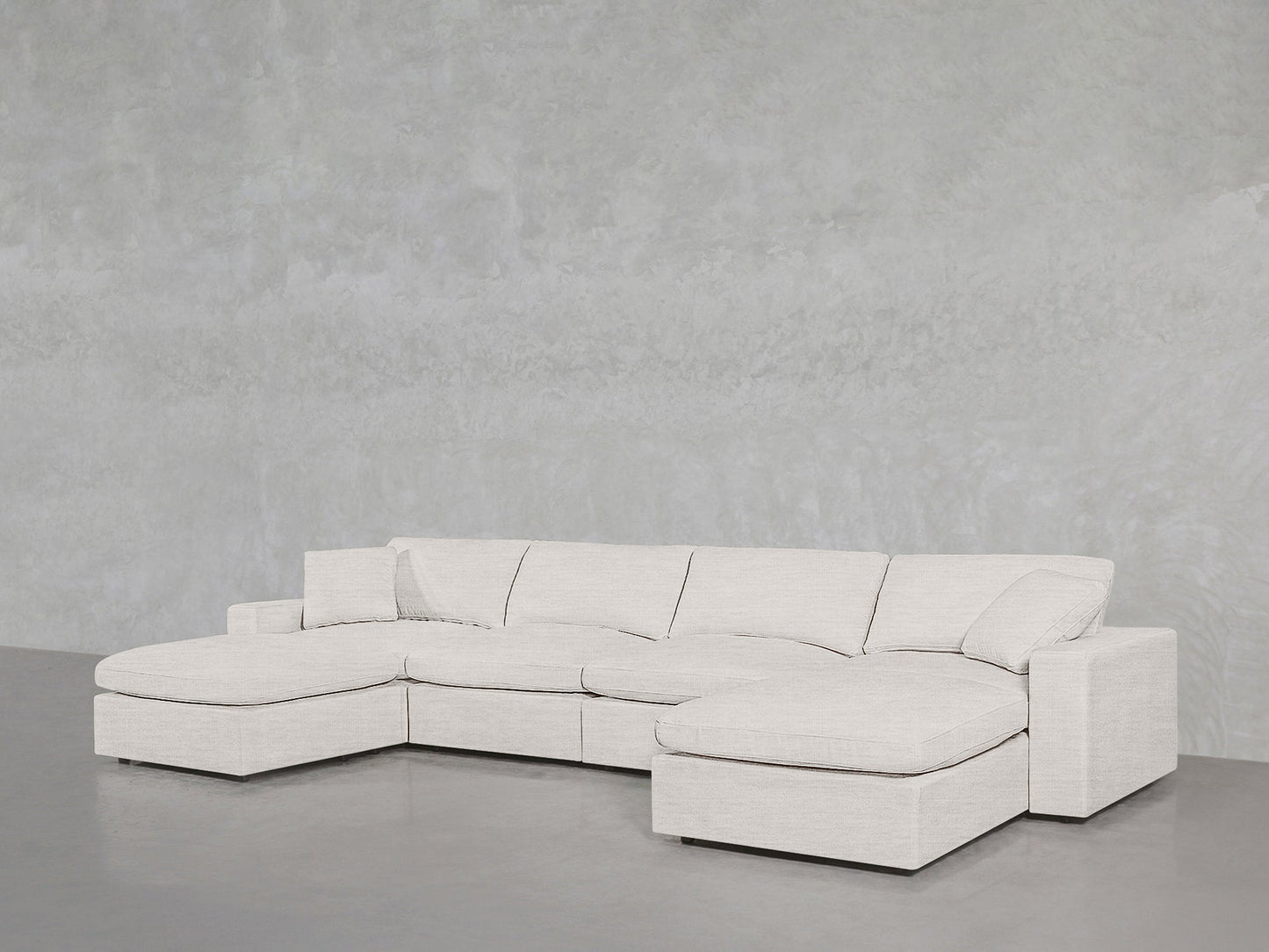 A spacious, modern 6-Seat Modular Double Chaise Sectional in light beige fabric is arranged against a textured light gray wall on a polished concrete floor.