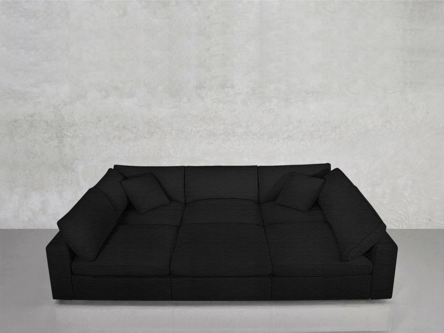 A 6-Seat Modular Pit in black with wide armrests and two matching cushions, set against a light grey textured wall on a smooth, light-colored floor.