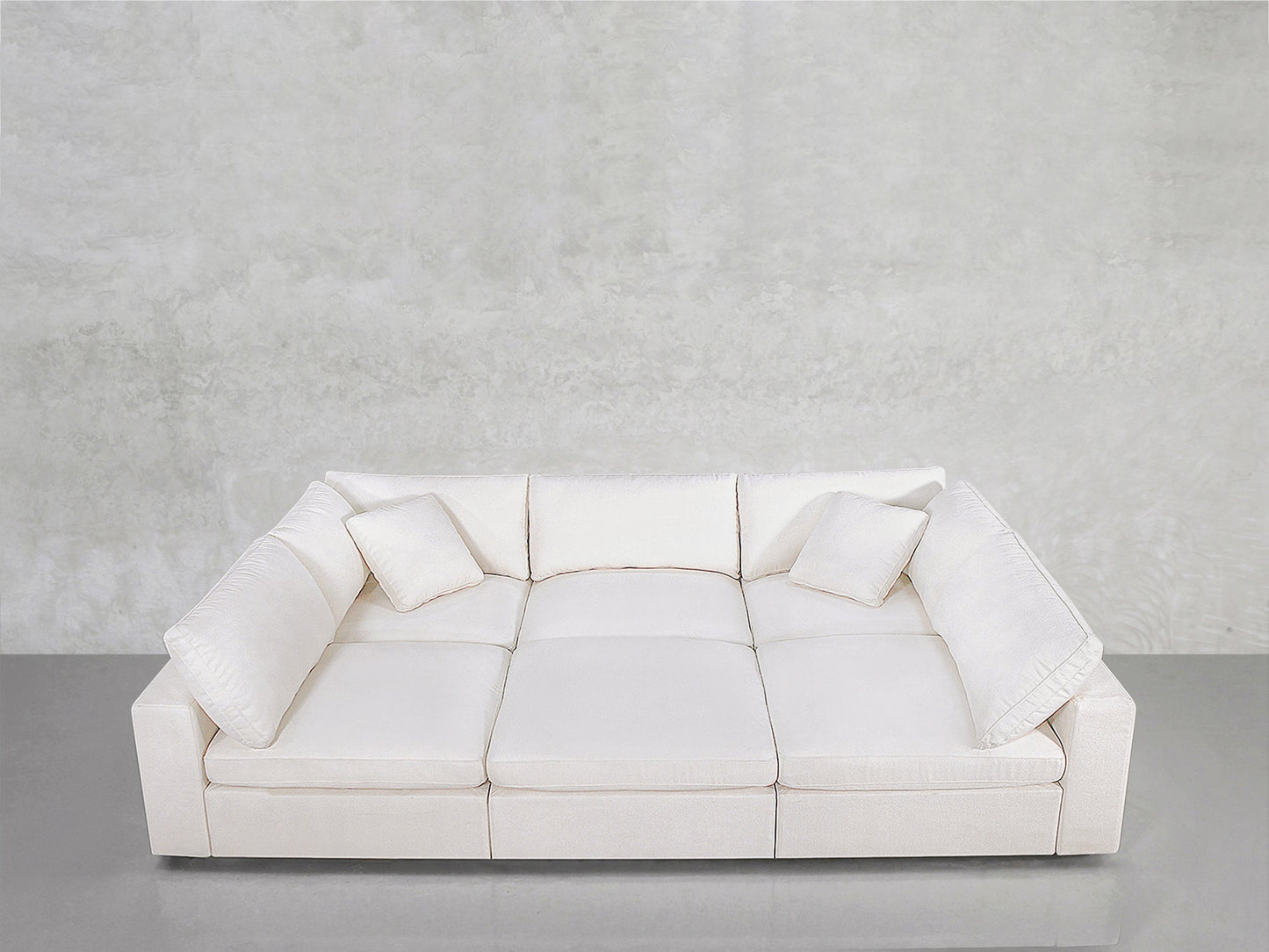 A contemporary white 6-Seat Modular Pit with three seat cushions and four matching throw pillows, positioned near a light gray textured wall and reflective gray floor.