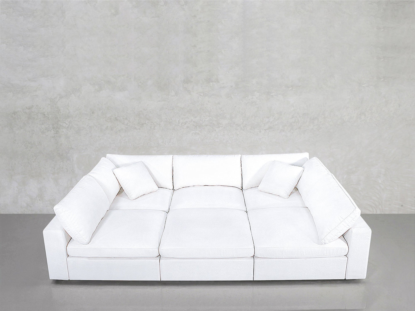 A modern white 6-Seat Modular Pit with three seat cushions and four throw pillows sits against a plain, light gray textured wall on a smooth gray floor.
