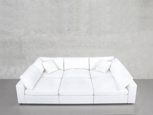 A modern white 6-Seat Modular Pit with three seat cushions and four throw pillows sits against a plain, light gray textured wall on a smooth gray floor.