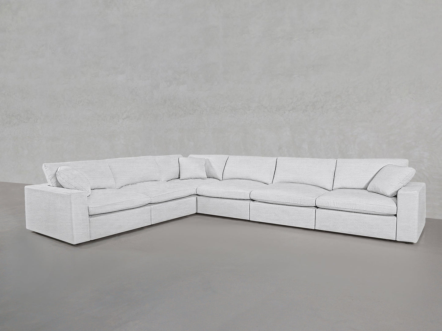 A 6-Seat Modular Corner Sectional with cushioned seats and wide armrests sits on a smooth gray floor, set against a plain light gray wall.