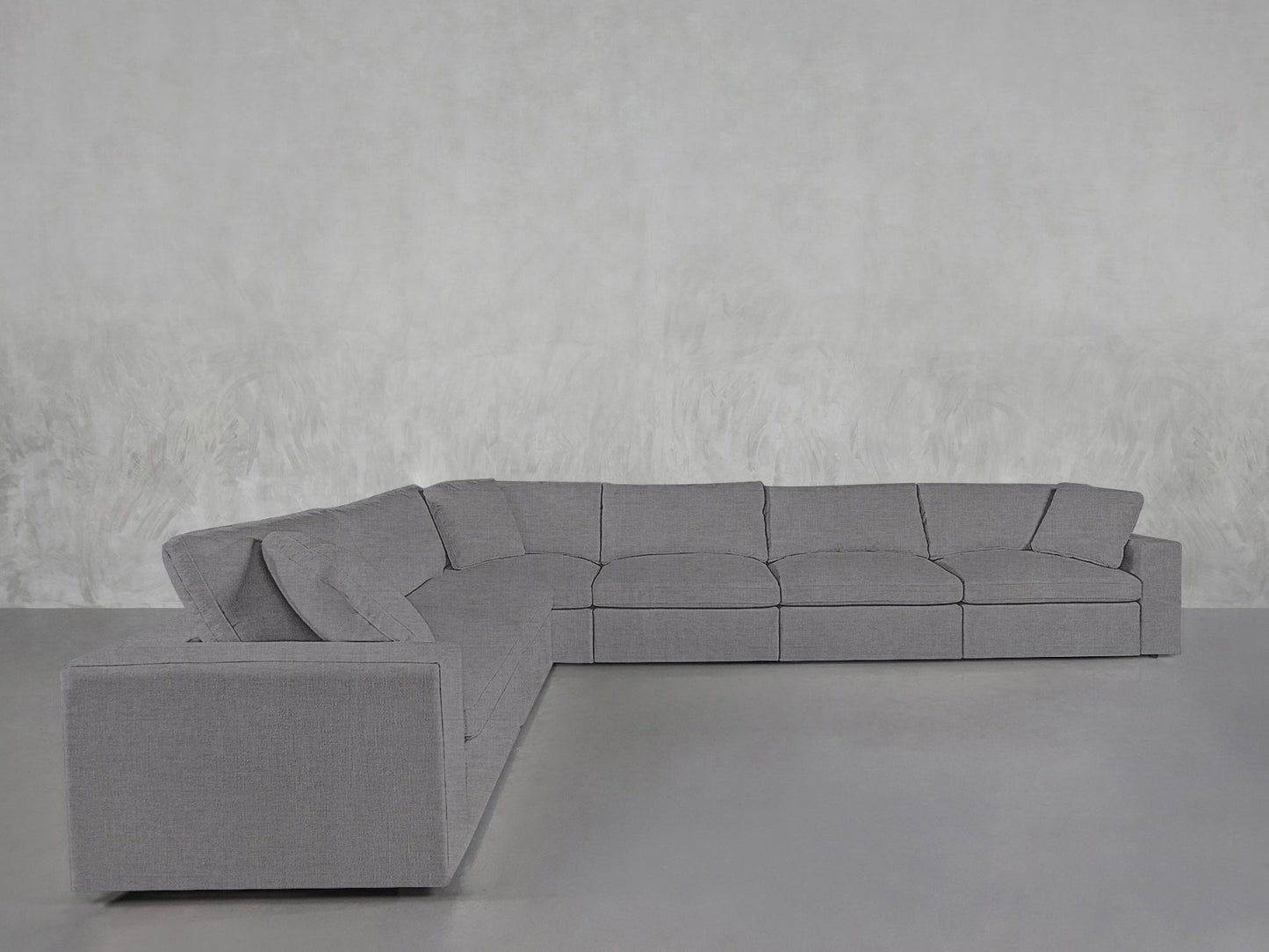 A 6-Seat Modular Corner Sectional with thick cushions and clean lines sits on a smooth gray floor against a light, textured wall, featuring a modern gray design and a left-facing chaise.