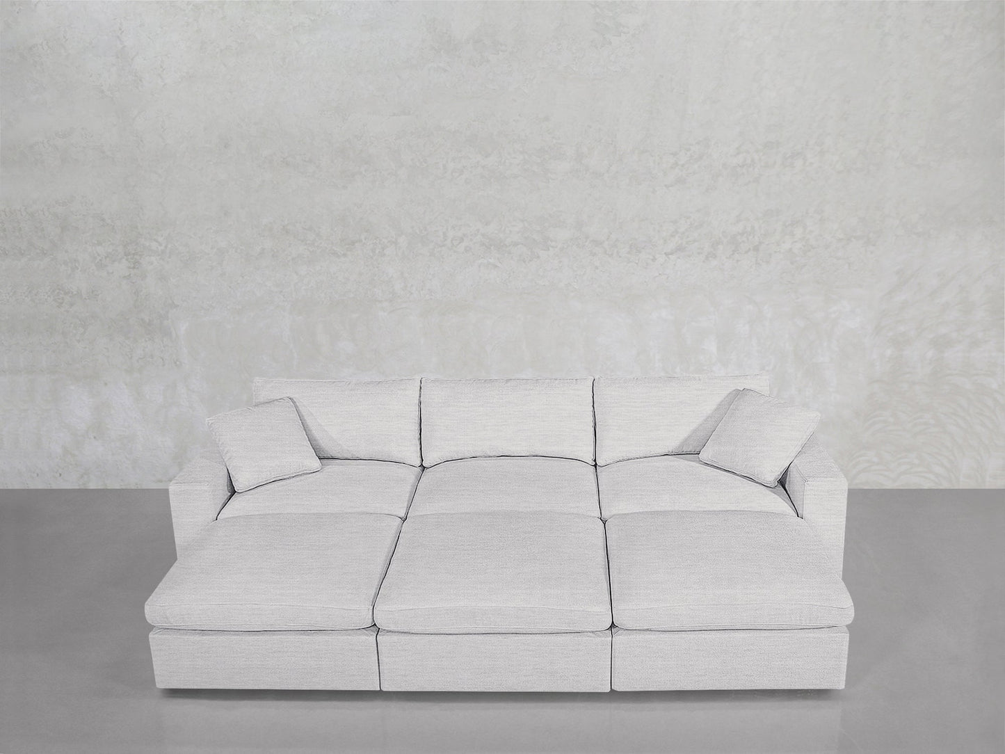 A modern 6-Seat Modular Daybed in light gray, featuring three seat cushions and two matching pillows, is set against a textured pale gray wall on a smooth gray floor.