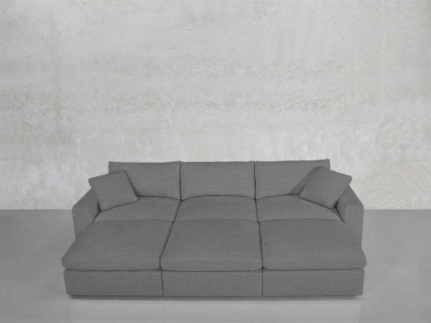 A contemporary 6-Seat Modular Daybed in gray, featuring three large seat cushions and four matching square back cushions, set against a plain light-textured wall on a smooth floor.