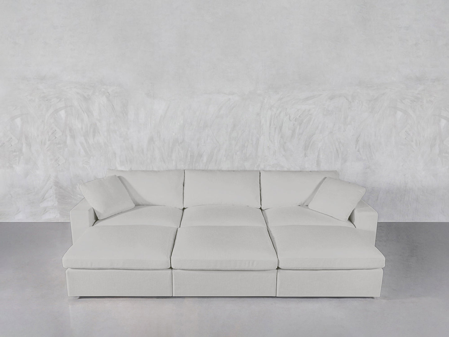 The 6-Seat Modular Daybed, in light gray with three large seat cushions and two pillows, is arranged against a textured white wall and smooth gray floor.