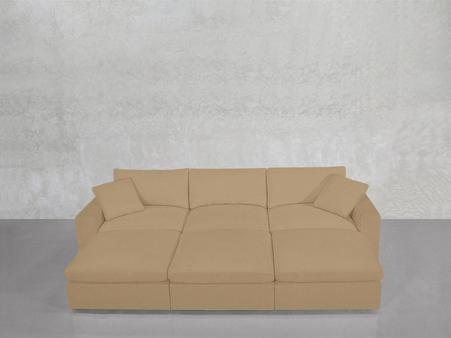 A 6-Seat Modular Daybed in beige, featuring three seat cushions and two matching throw pillows, is arranged against a light gray textured wall and a smooth gray floor.