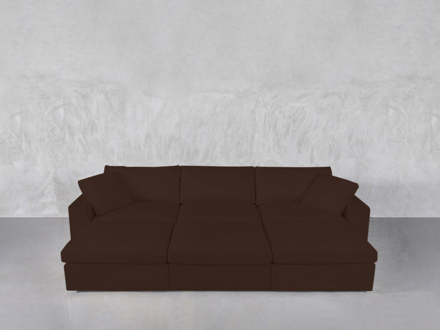 A 6-Seat Modular Daybed in dark brown, featuring three seat cushions and three back cushions, set against a plain light gray textured wall and a light gray floor.