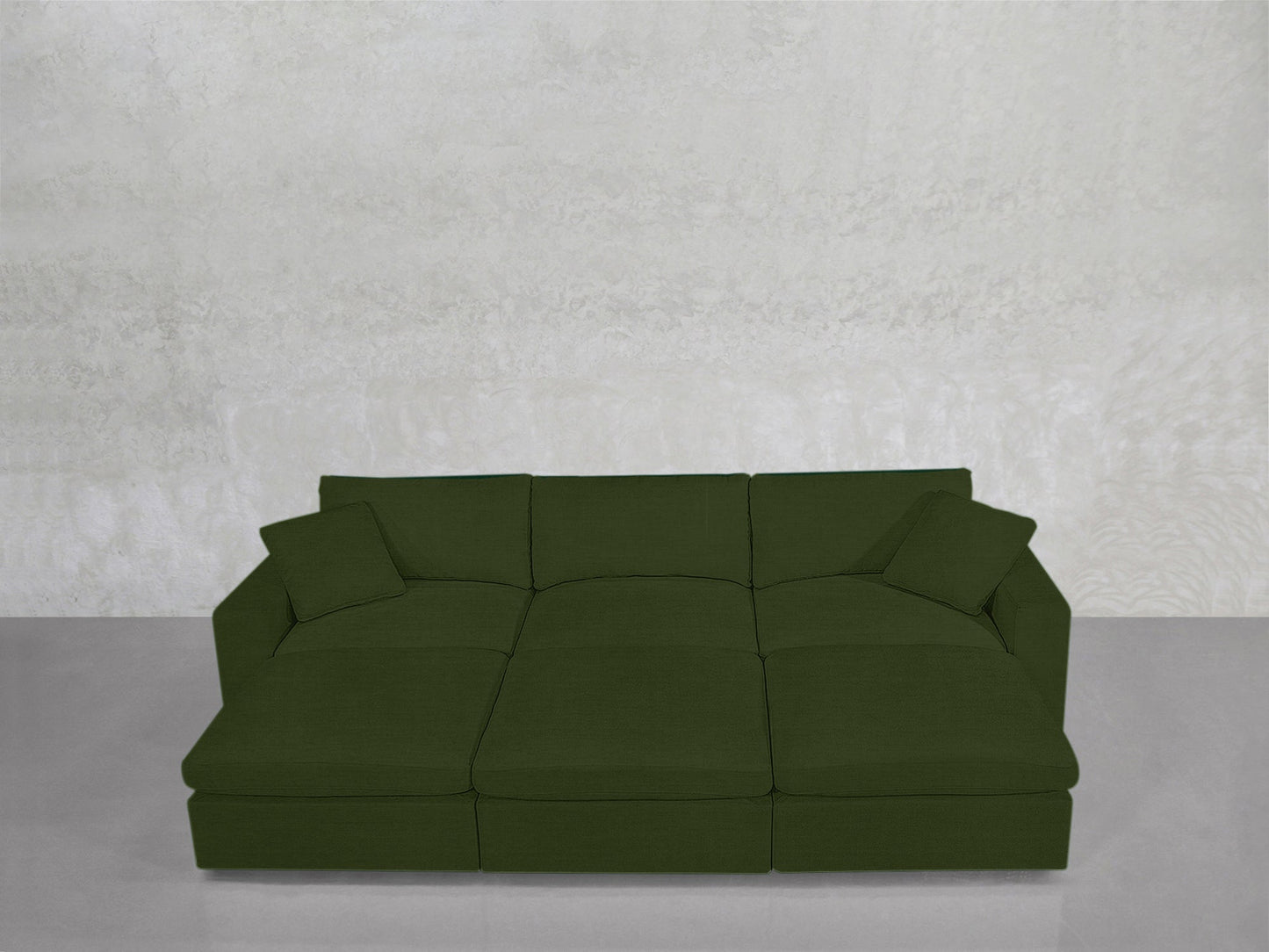 A 6-Seat Modular Daybed in deep green with wide seats and two matching square pillows sits against a light textured wall on a smooth, light gray floor.