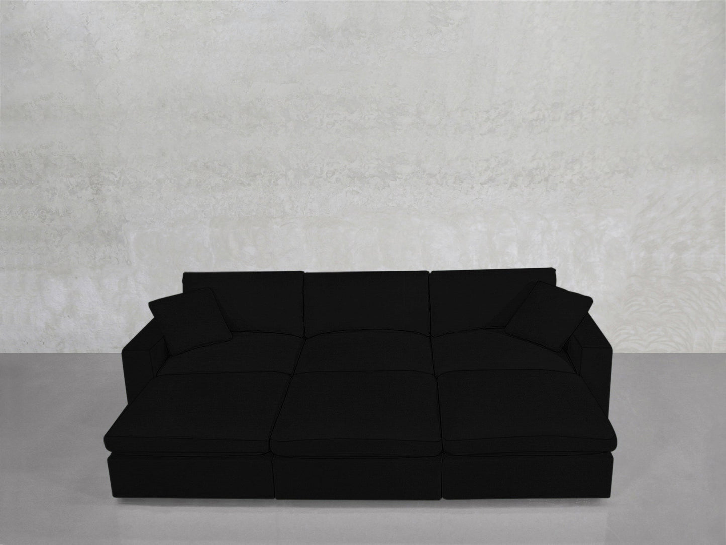The 6-Seat Modular Daybed in black, featuring two matching cushions, is showcased against a light gray textured wall and a smooth, light gray floor.