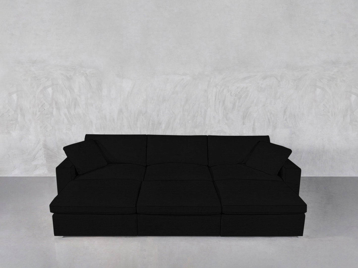 A modern black 6-seat modular daybed with two matching black throw pillows sits against a textured light gray wall on a polished gray floor.