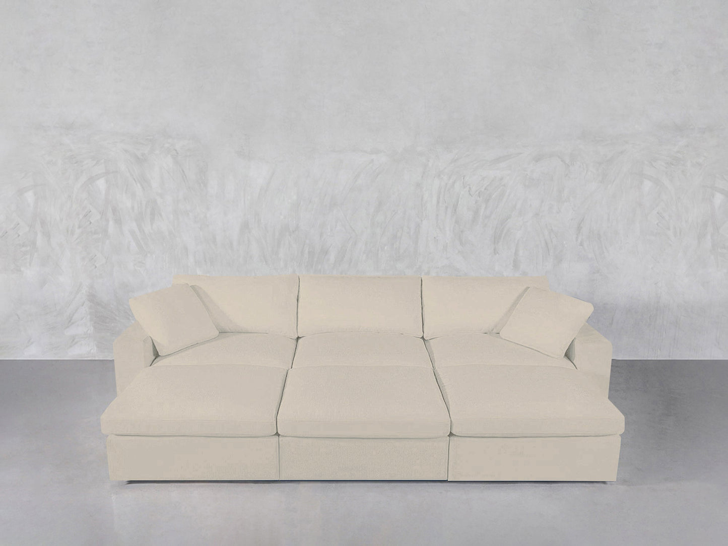 A beige, modern 6-Seat Modular Daybed with two matching throw pillows is placed against a textured light gray wall and a smooth gray floor.