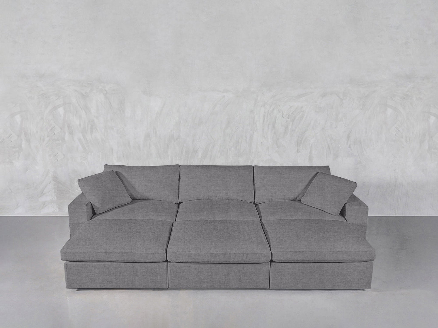 The 6-Seat Modular Daybed, featuring three seat cushions and four matching pillows, sits against a light gray textured wall on a smooth, light-colored floor.