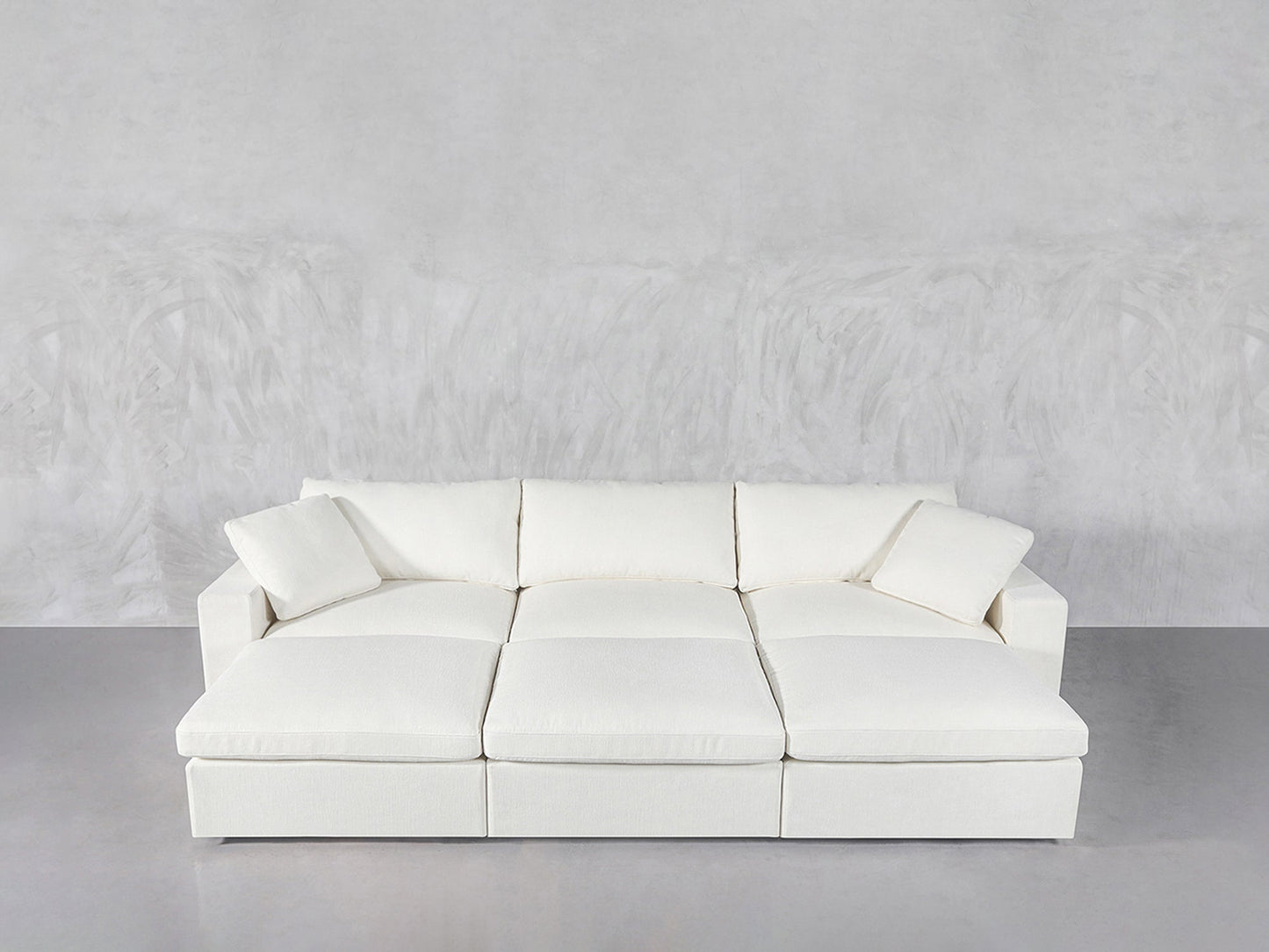 A modern, white 6-Seat Modular Daybed with wide cushions and two matching throw pillows, set against a light gray textured wall and floor.