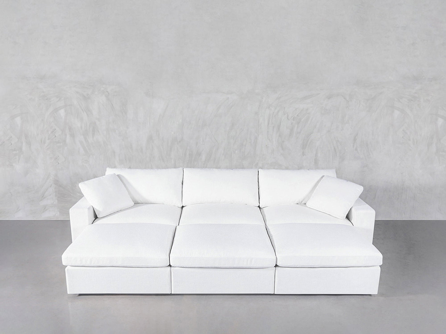 A 6-Seat Modular Daybed in modern white with three seat cushions and two matching throw pillows sits against a light gray textured wall on a smooth gray floor.