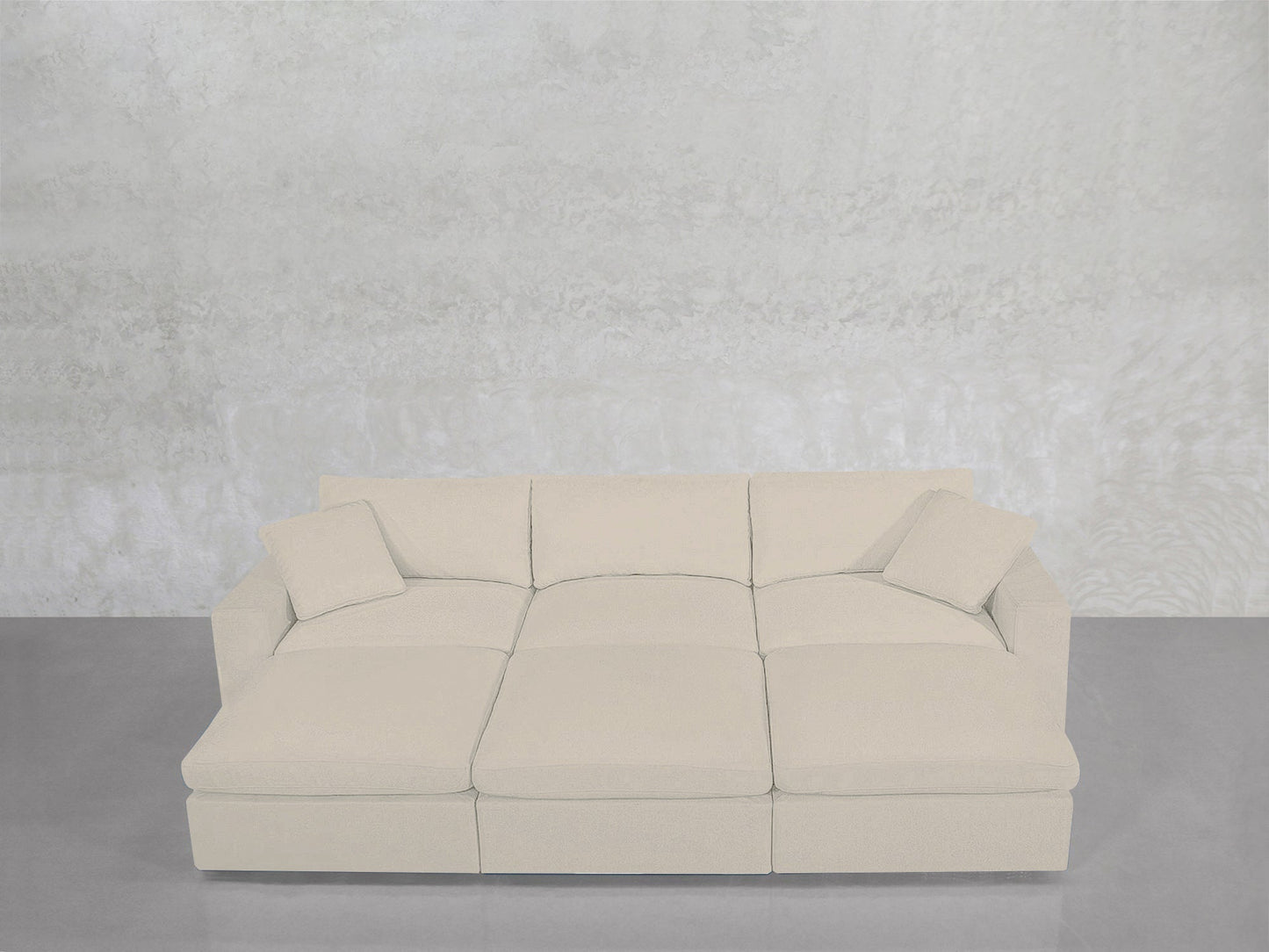 A modern light beige 6-Seat Modular Daybed with matching square pillows is set against a textured light gray wall on a smooth gray floor.