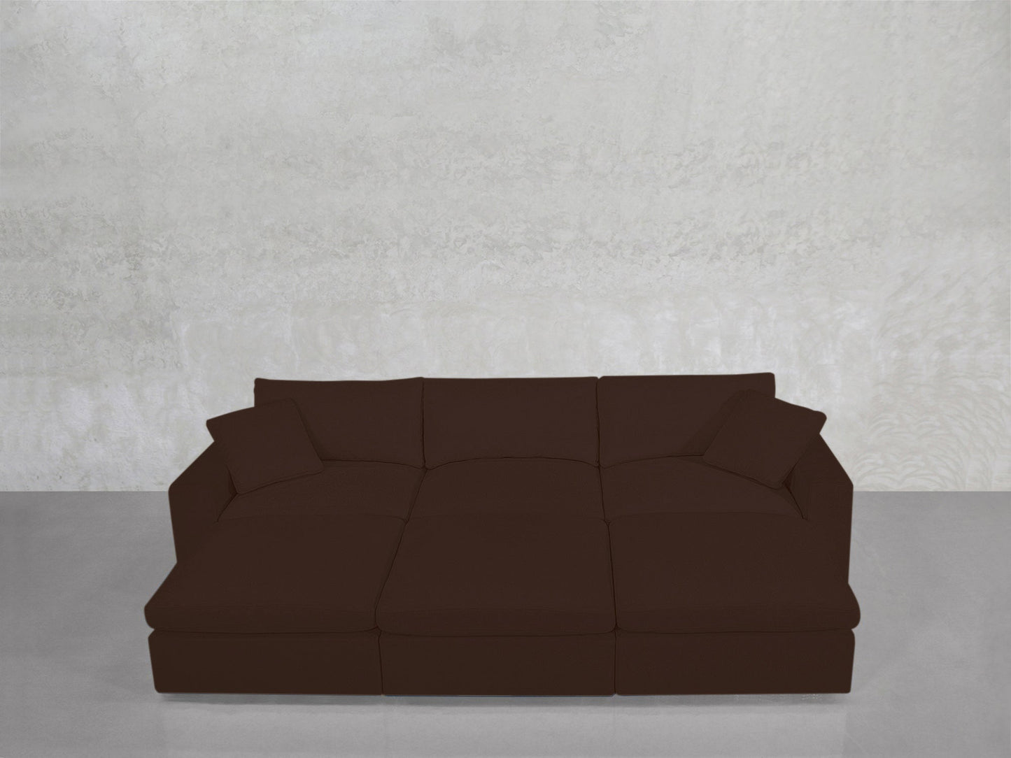 A dark brown 6-Seat Modular Daybed with wide armrests and two matching cushions sits against a light gray textured wall on a polished gray floor.