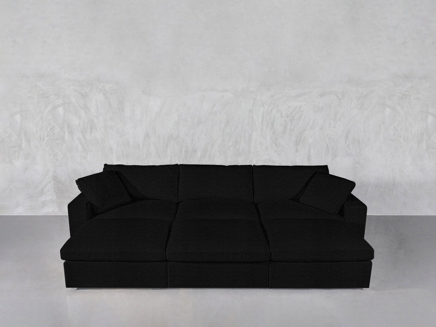 A modern black 6-Seat Modular Daybed with three seat cushions, three back cushions, and two matching throw pillows is placed against a light gray textured wall and floor.