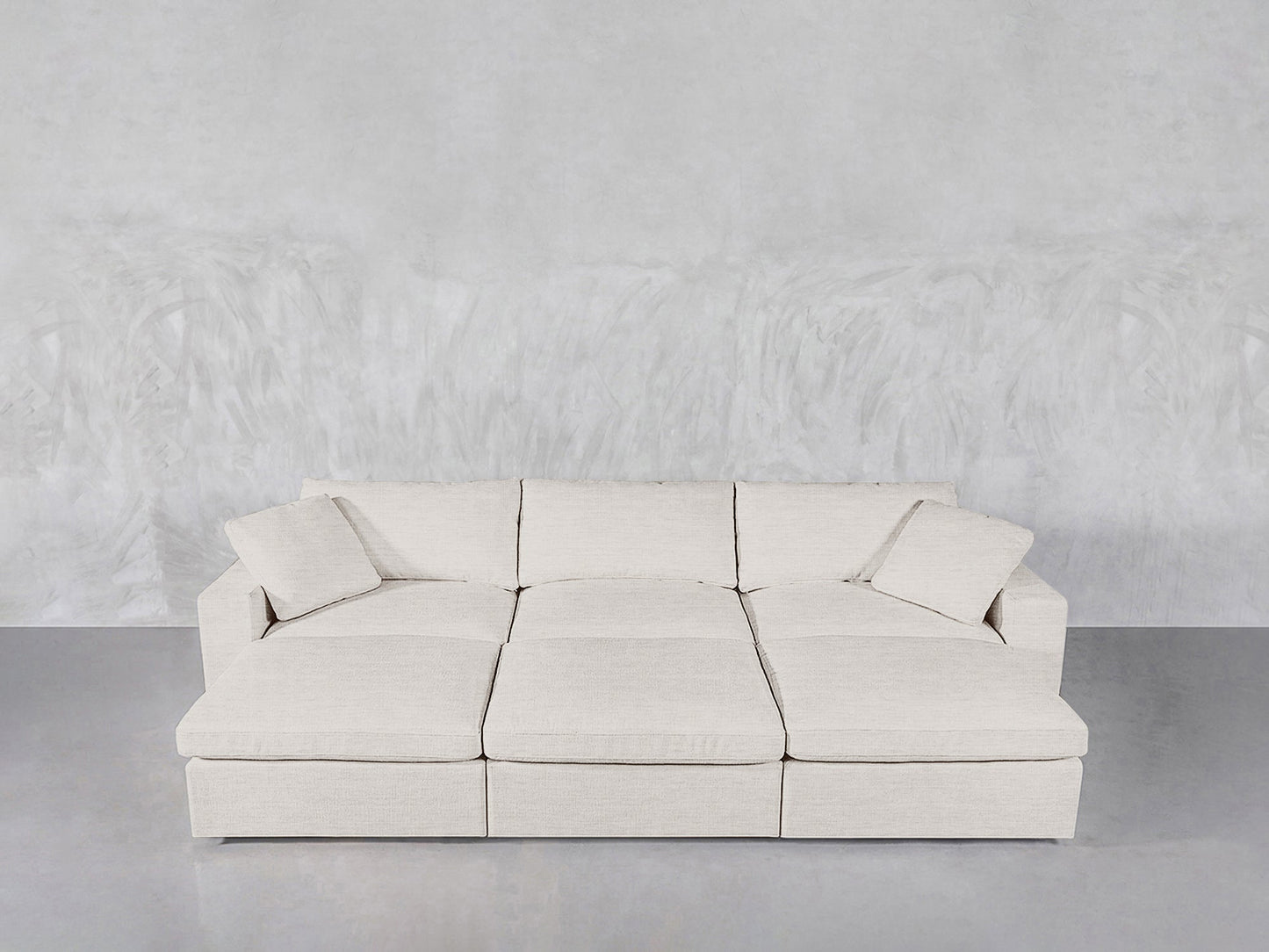 A 6-Seat Modular Daybed in light beige with wide seats and two matching cushions is placed against a textured light gray wall on a smooth gray floor.