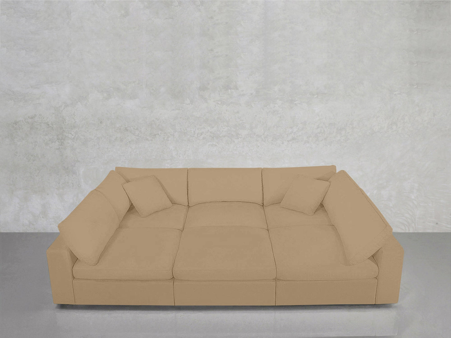 A 6-Seat Modular Pit in beige, featuring three seat cushions and four matching throw pillows, is placed against a textured light gray wall on a shiny gray floor.