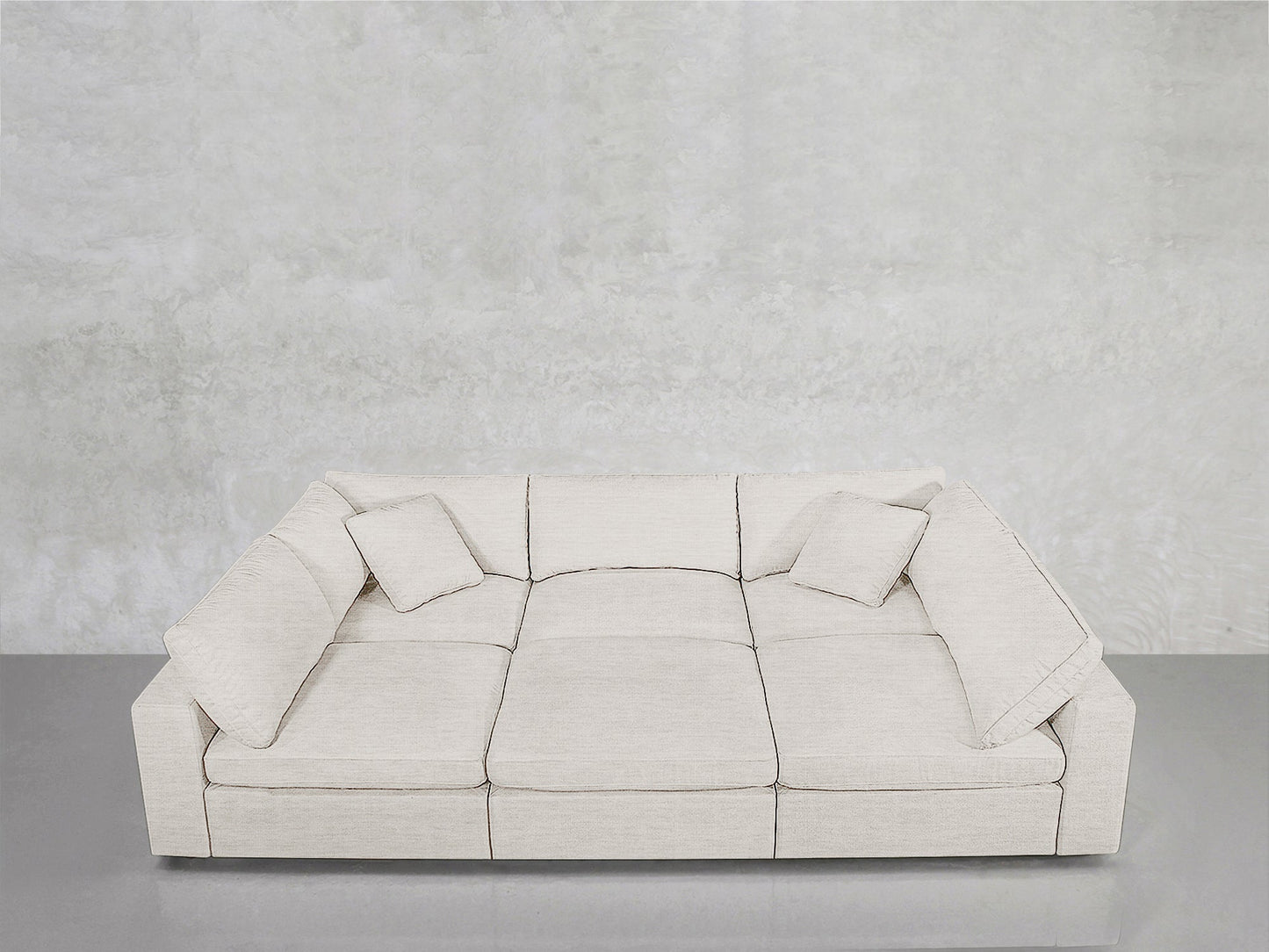 A contemporary 6-Seat Modular Pit in light beige with three seat cushions and two matching pillows sits on a shiny gray floor against a textured light gray wall.