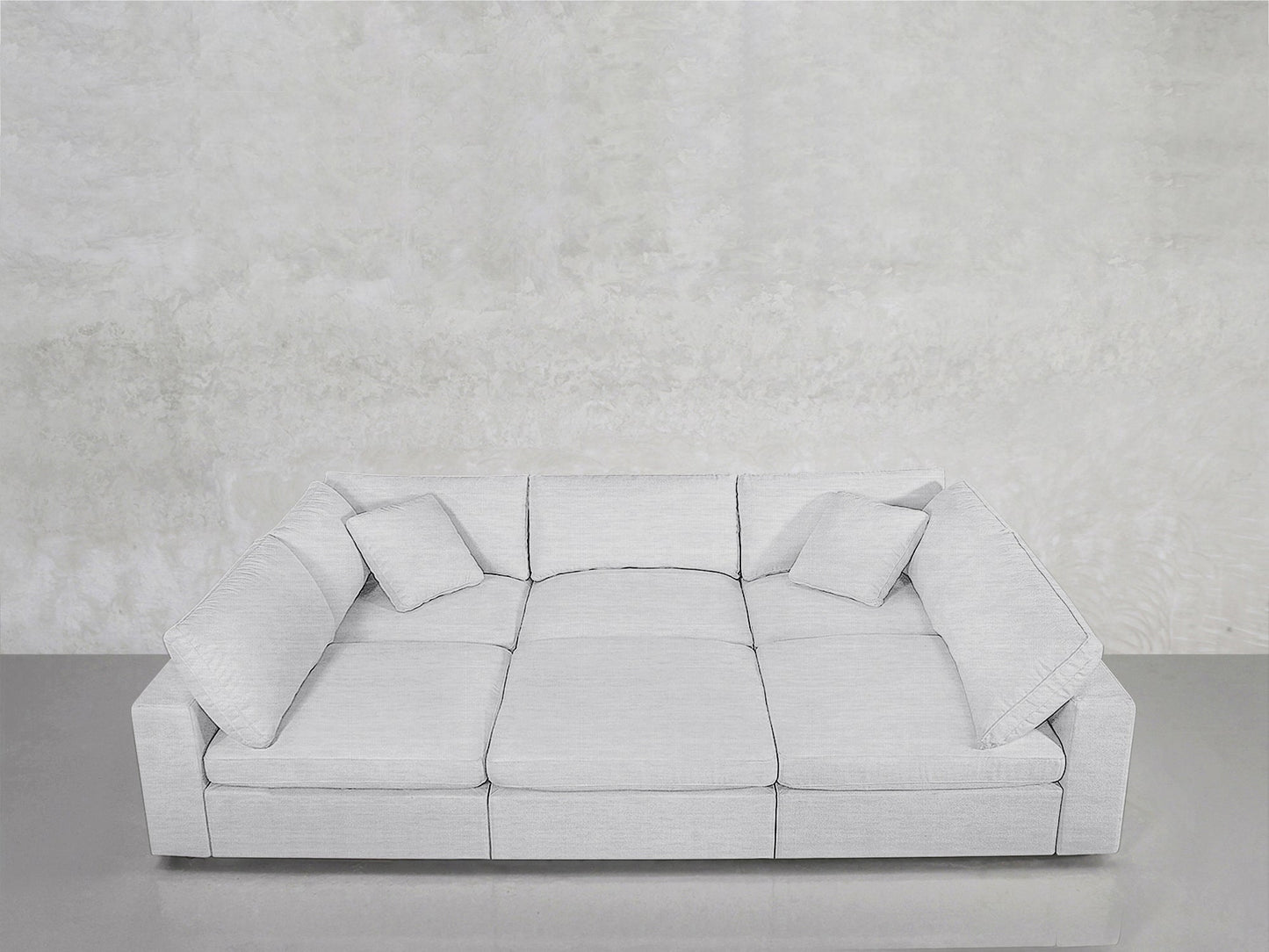 A modern 6-Seat Modular Pit in light gray, featuring three seat cushions and four matching throw pillows, is set against a plain, textured white wall on a smooth gray floor.