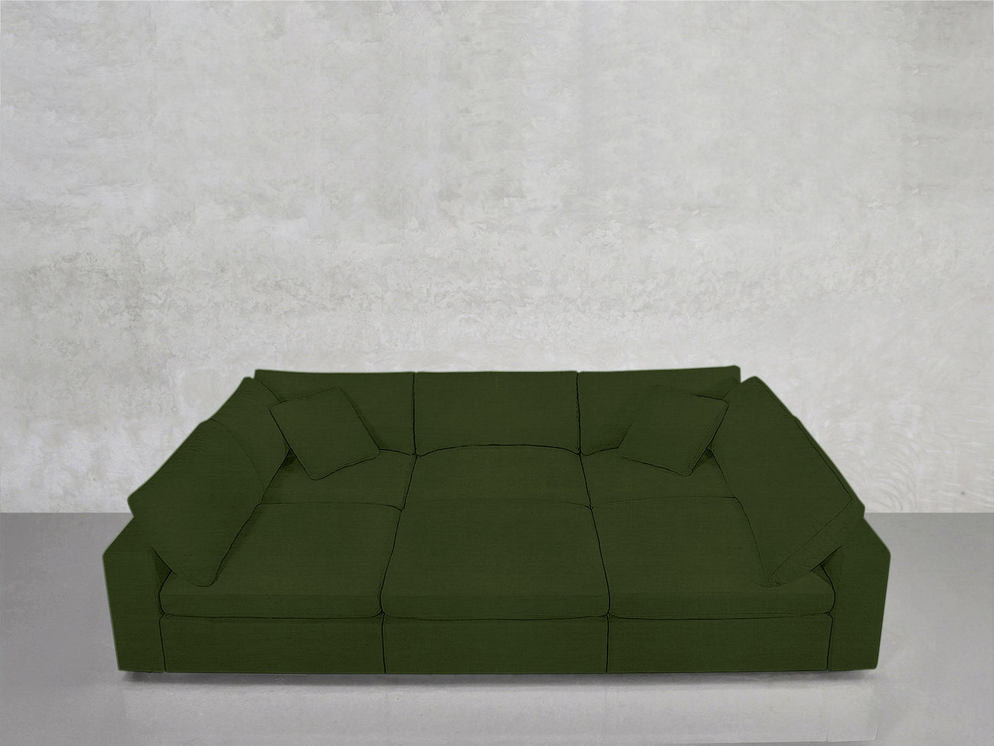 The 6-Seat Modular Pit, a large deep green sectional with three seat cushions and matching pillows, sits against a textured light gray wall on a smooth, light gray floor.