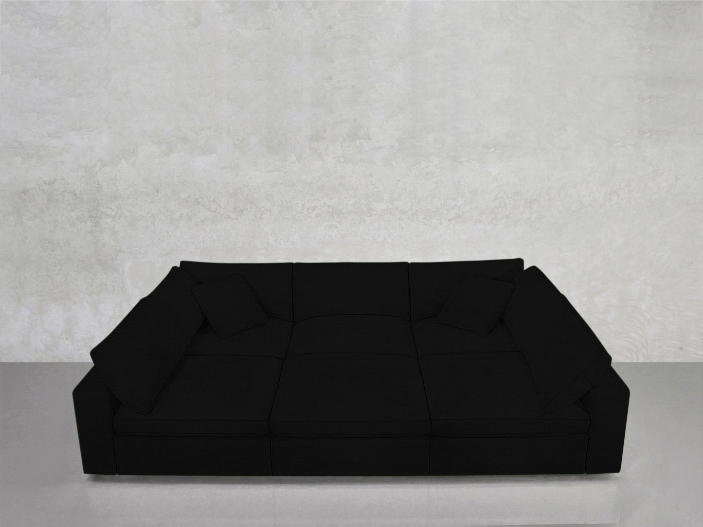 The 6-Seat Modular Pit, a spacious modern black sofa with wide armrests and four cushions, sits against a light gray textured wall on a smooth, reflective floor.
