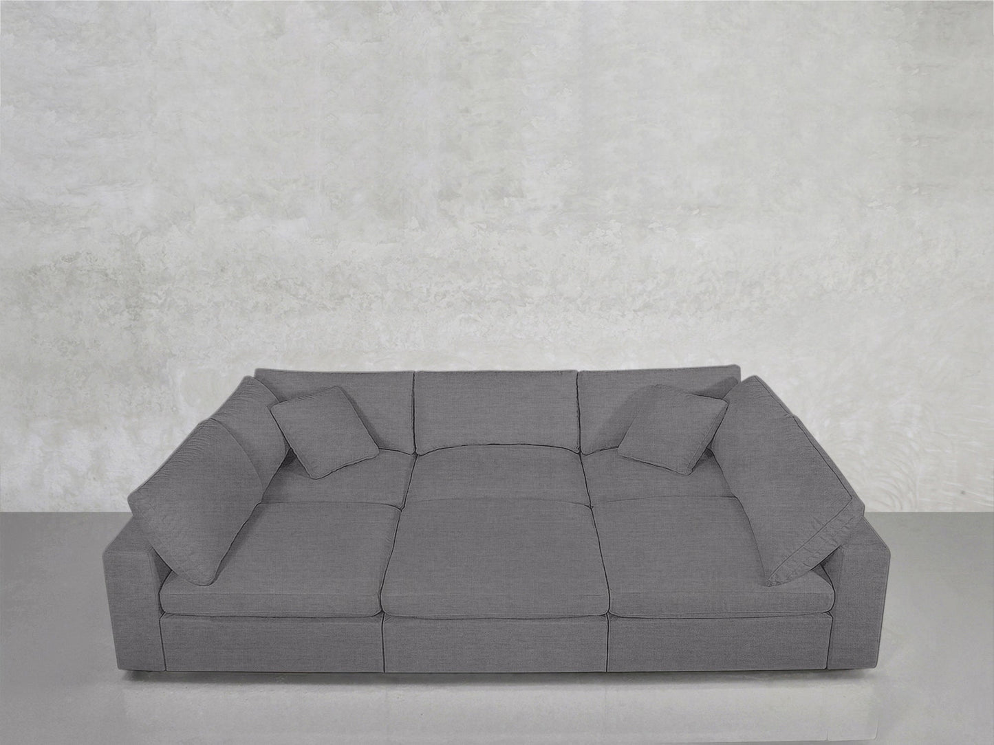 The 6-Seat Modular Pit, a spacious modern gray sofa with squared cushions and three matching throw pillows, sits against a light gray textured wall and floor.