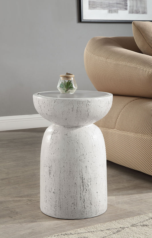 The Dylana Natural Fiber Concrete Accent Table in gray stands by a beige sofa, topped with a small glass terrarium holding a plant, set against a gray wall with framed artwork.