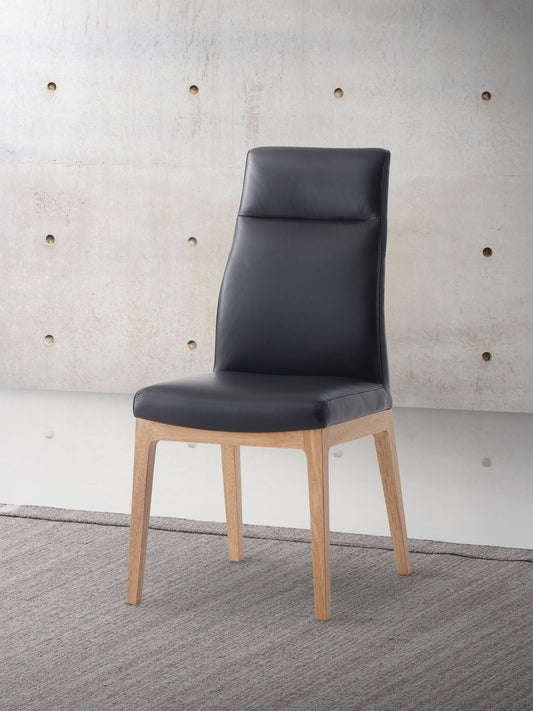 The Raquan - Leather Side Chair (Set of 2) - Black Pu features a black cushioned seat and back, light wood legs, and is shown on a gray rug before a beige concrete wall with circular indentations.