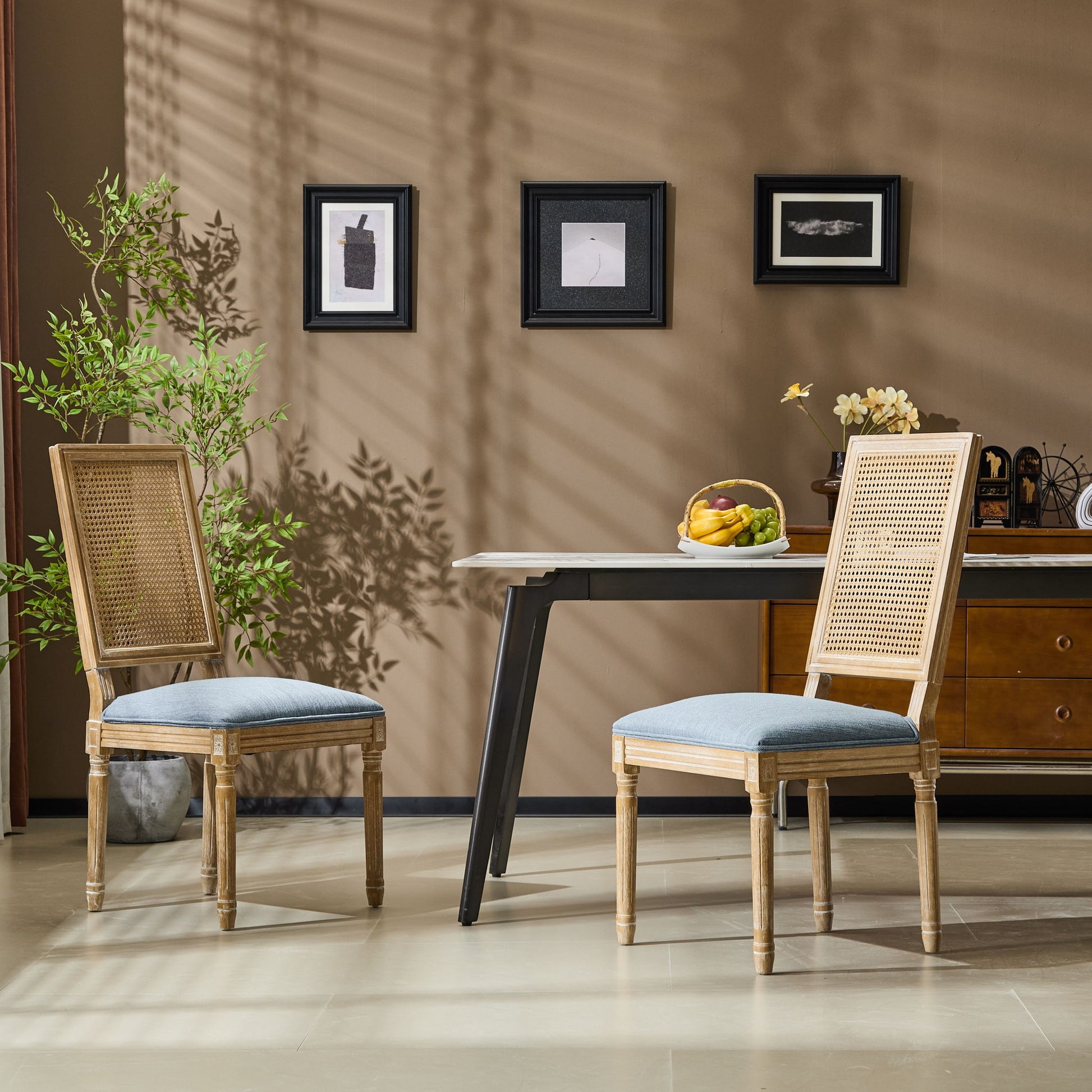 Two French Country Dining Chairs with rectangular cane backrests and smooth upholstered seats in light blue sit by a modern table, topped with a fruit bowl, near a brown wall with framed art and shadows from a leafy plant.