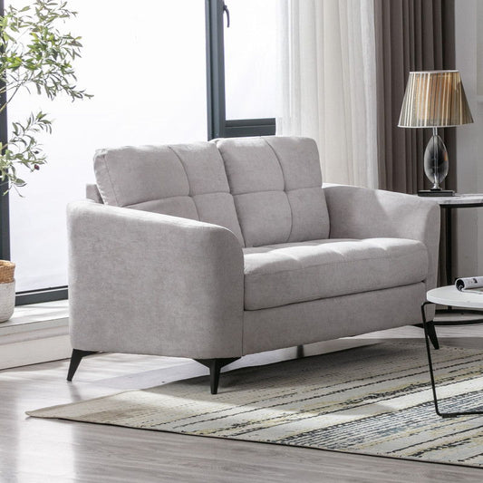 The Callie Woven Fabric Loveseat in light gray with tufted cushions is placed in a bright living room by a window, accompanied by a small round side table with a lamp and a striped rug on a wooden floor.
