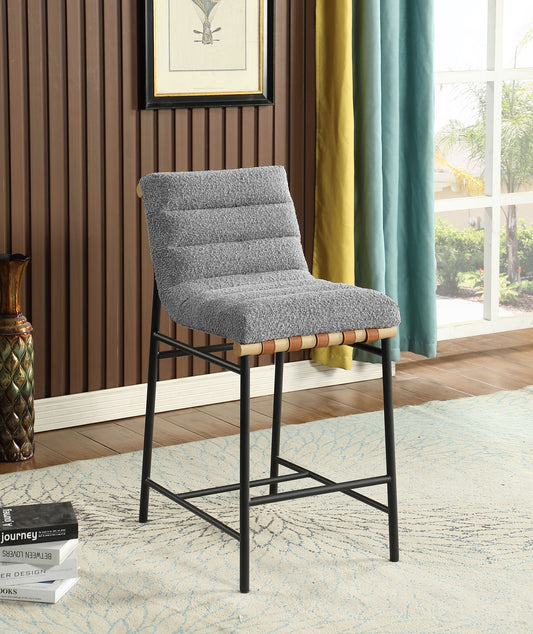 The Lahni - 17" Boucle Fabric Counter Height Chair, featuring a gray cushioned seat and black metal legs, stands on a light patterned rug in a wood-paneled room with large windows, colorful curtains, and framed artwork on the wall.
