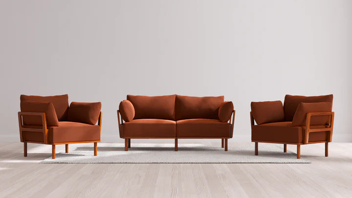 The Indoor Jack 2-Seater Sofa + Armchair Set in Balsa, with its rust-colored upholstery and wooden frames, is elegantly arranged on a light gray area rug. The setting features a plain, light-colored wall and a wooden floor in the background.