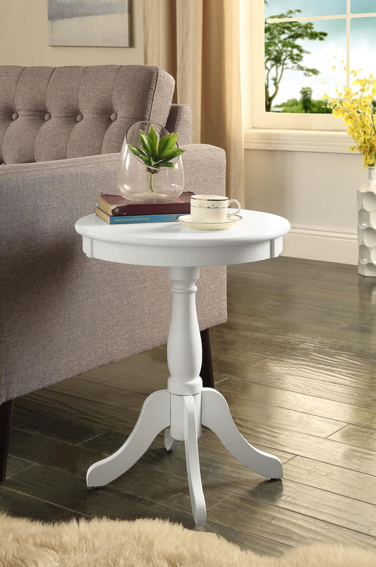 The Alger Accent Table in white sits by a gray sofa, holding a glass vase with a plant, a book, and a teacup. Behind it, wooden flooring leads to a window with curtains and yellow flowers in the background.