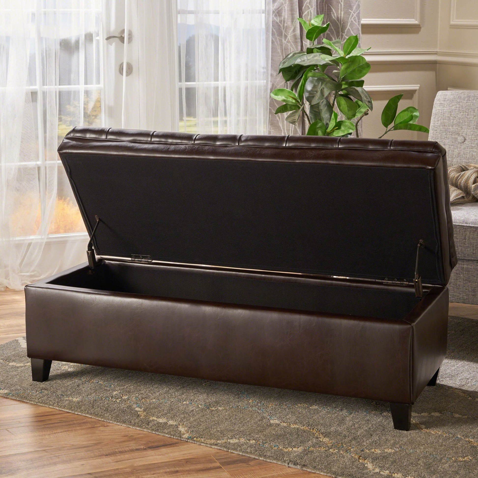 The Juliana Storage Ottoman with Birch Legs, in dark brown leather with an open lid, sits on a gray rug in a bright room featuring large windows, white curtains, and a potted plant nearby.