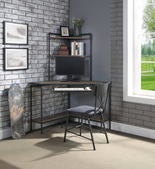 A modern corner desk setup with a computer monitor, keyboard, books, and the Brantley - Office Chair in Antique Oak / Sandy Gray. Gray brick walls feature framed pictures and a skateboard. A large window reveals greenery outside.