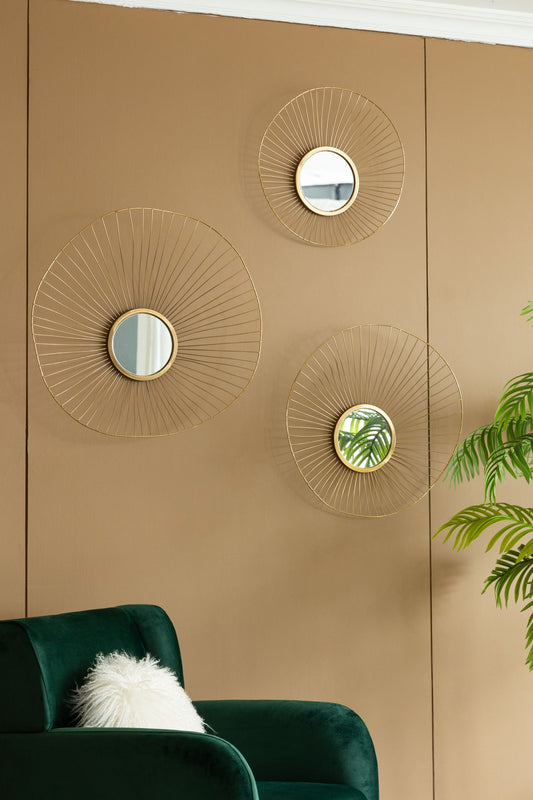 A tan wall displays the Wall Mirror Abstract Designed Wall Mirrors With Frame For Home & Office, Top Of Sideboard (Set of 3) - Gold. Below is a green velvet chair with a white fluffy pillow, next to a leafy green plant.