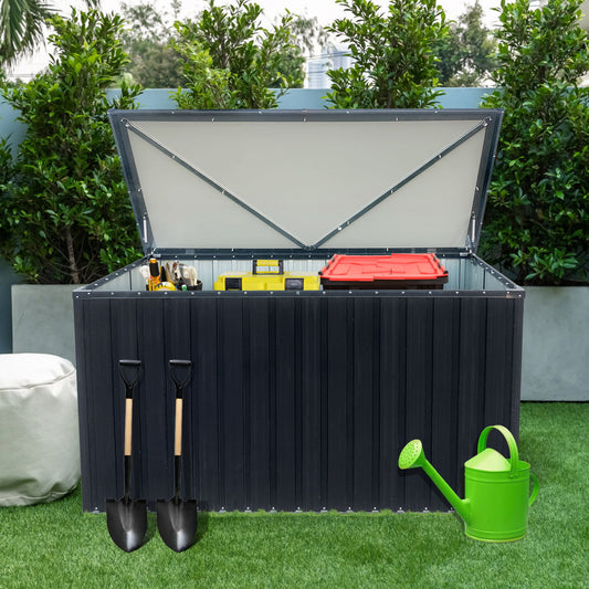 The Out Door Metal Storage Box Store Medium - Black is open, displaying gardening tools and a red container inside. Two shovels hang on the front, while a green watering can rests on artificial grass with green bushes in the background.