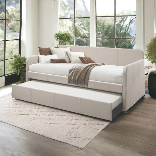 Modern living room with large windows, the Jagger Twin Daybed With Trundle in Fog, assorted throw pillows, a brown throw blanket, potted plants, and a textured rug on wood flooring. Sunlight filters through the trees outside.