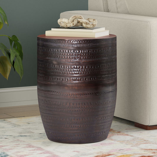 The Johnsen - Handcrafted Large Metal Accent Table, featuring dark bronze finish and textured horizontal patterns, stands beside a beige sofa. On top are stacked books and a white decor piece; nearby, a plant and patterned rug are partially visible.
