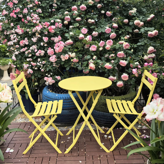 The 3 Piece Patio Bistro Set of a foldable round table and two chairs sits on a brick patio, surrounded by blooming pink roses and lush greenery.