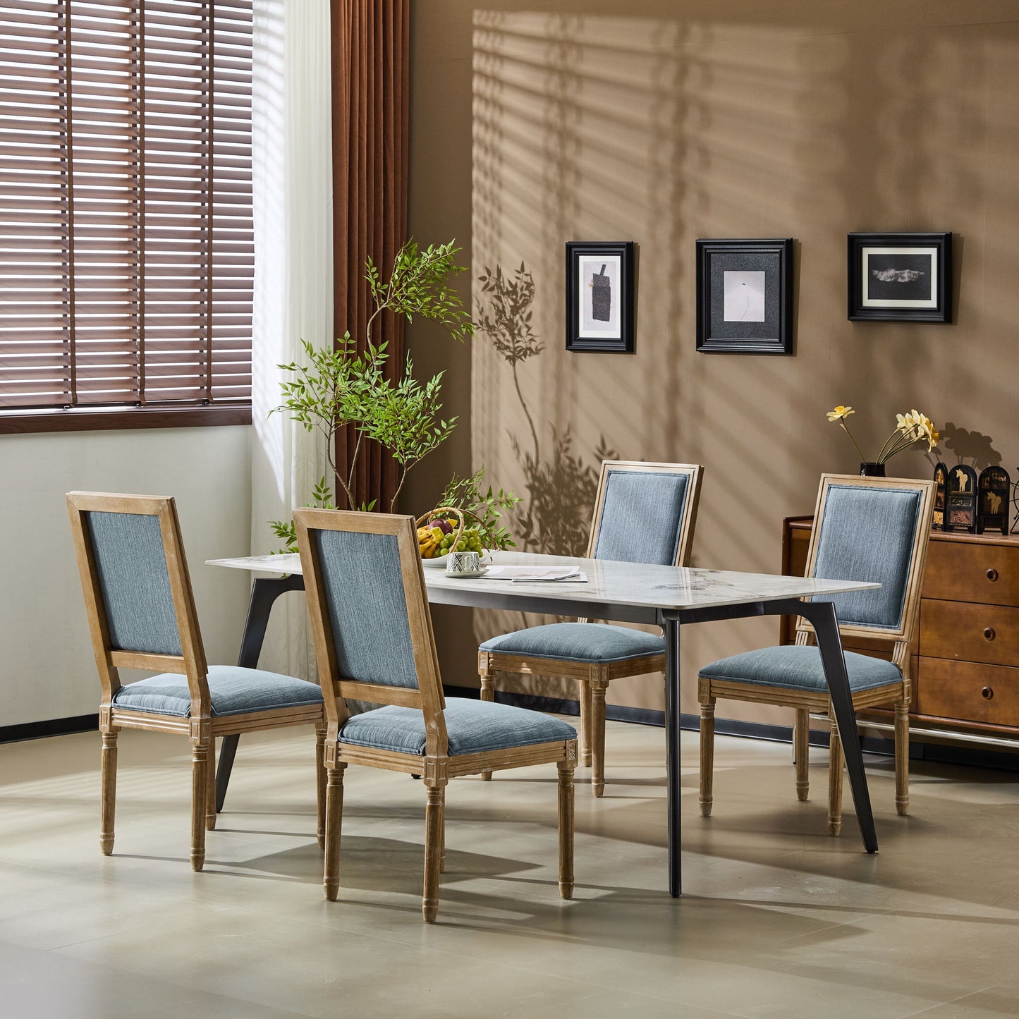 A dining room features a rectangular table, four French Country Style Dining Chairs with upholstered seats and fluted legs, a plant, and three framed pictures on a beige wall. Sunlight streams through window blinds on the left.